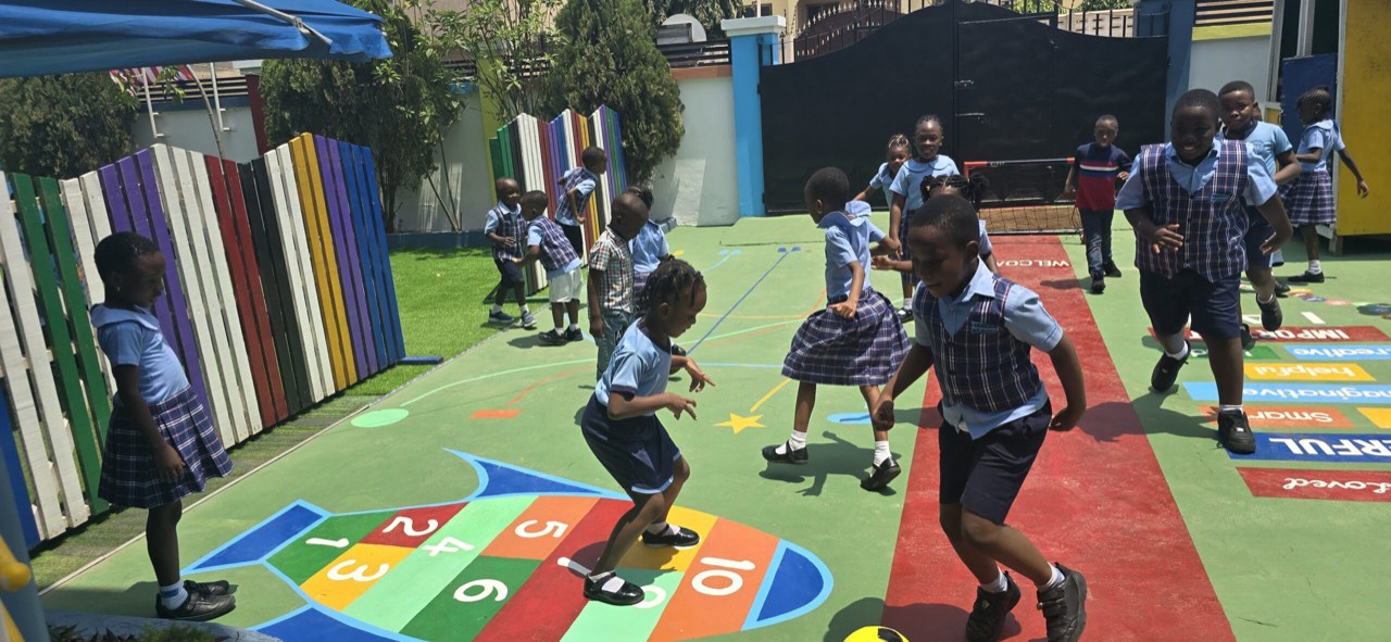 Pupils in blue Janique uniform playing on a green playground painted with a hopscotch grid, beside a rainbow-coloured fence