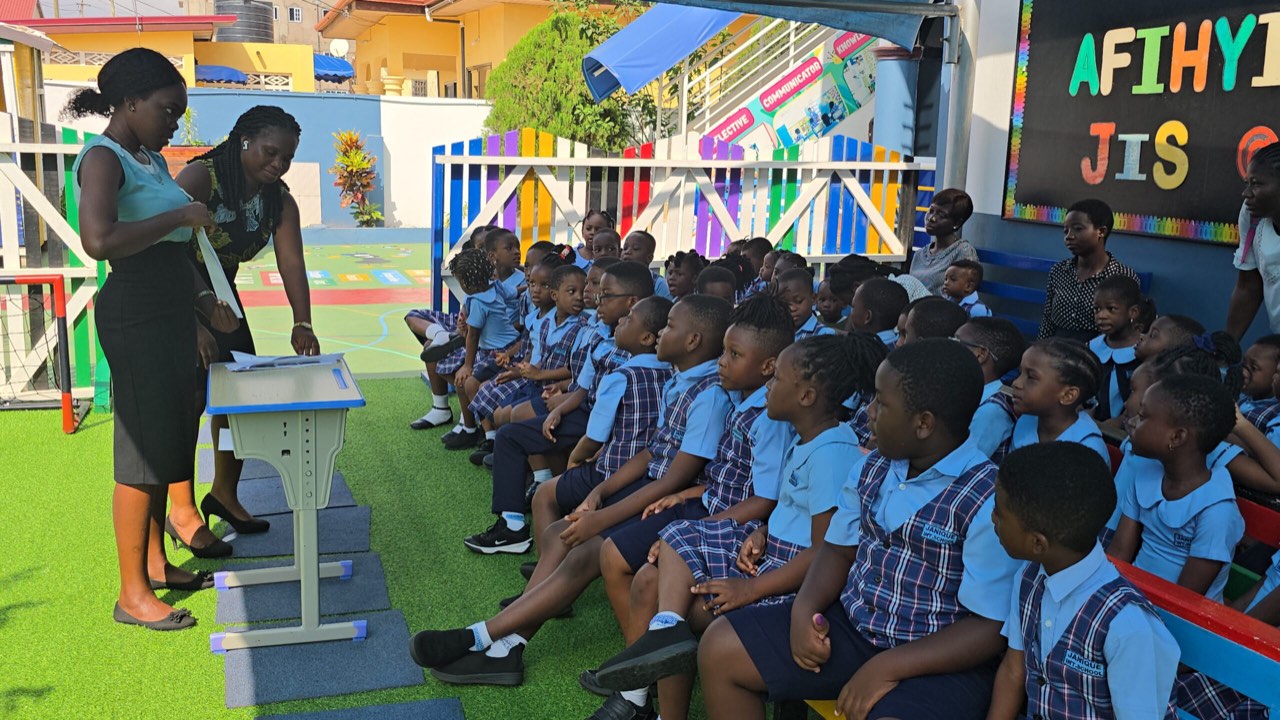 Rows of pupils seated outdoors watching two teachers at a lectern, in front of a lettered display board